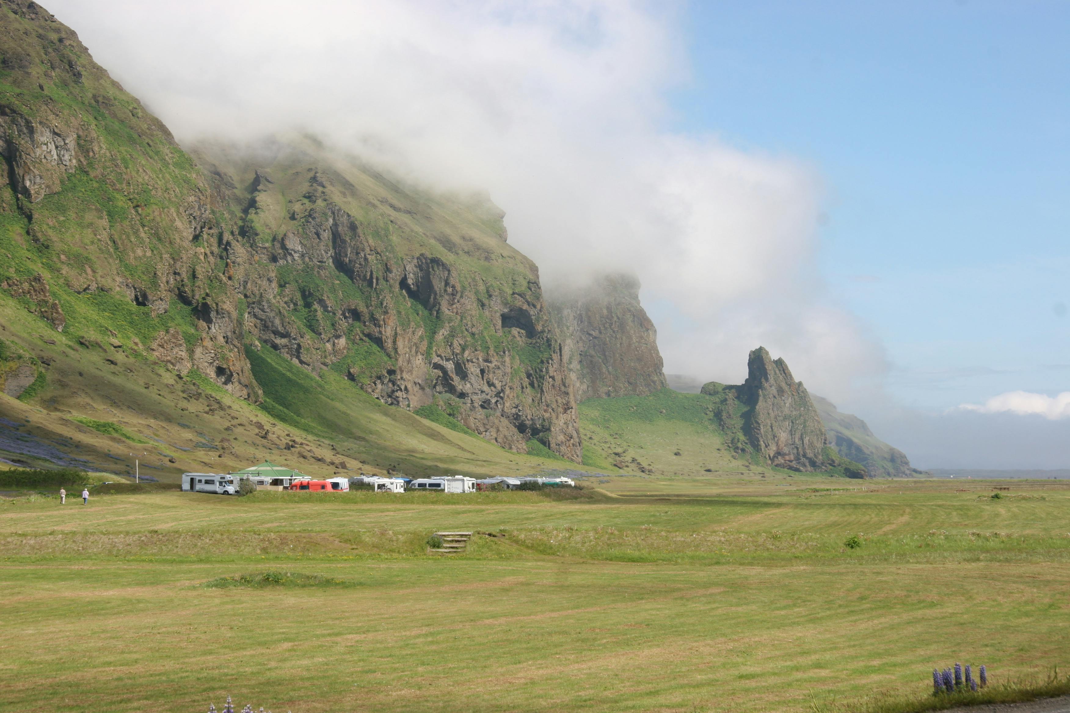 A grassy field with scattered RVs and trailers, bordered by green, rocky mountains partially covered in low-hanging clouds.
