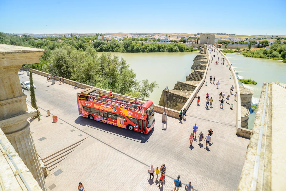 A red double-decker tour bus near a riverside stone bridge with people walking and lush greenery in the background.
