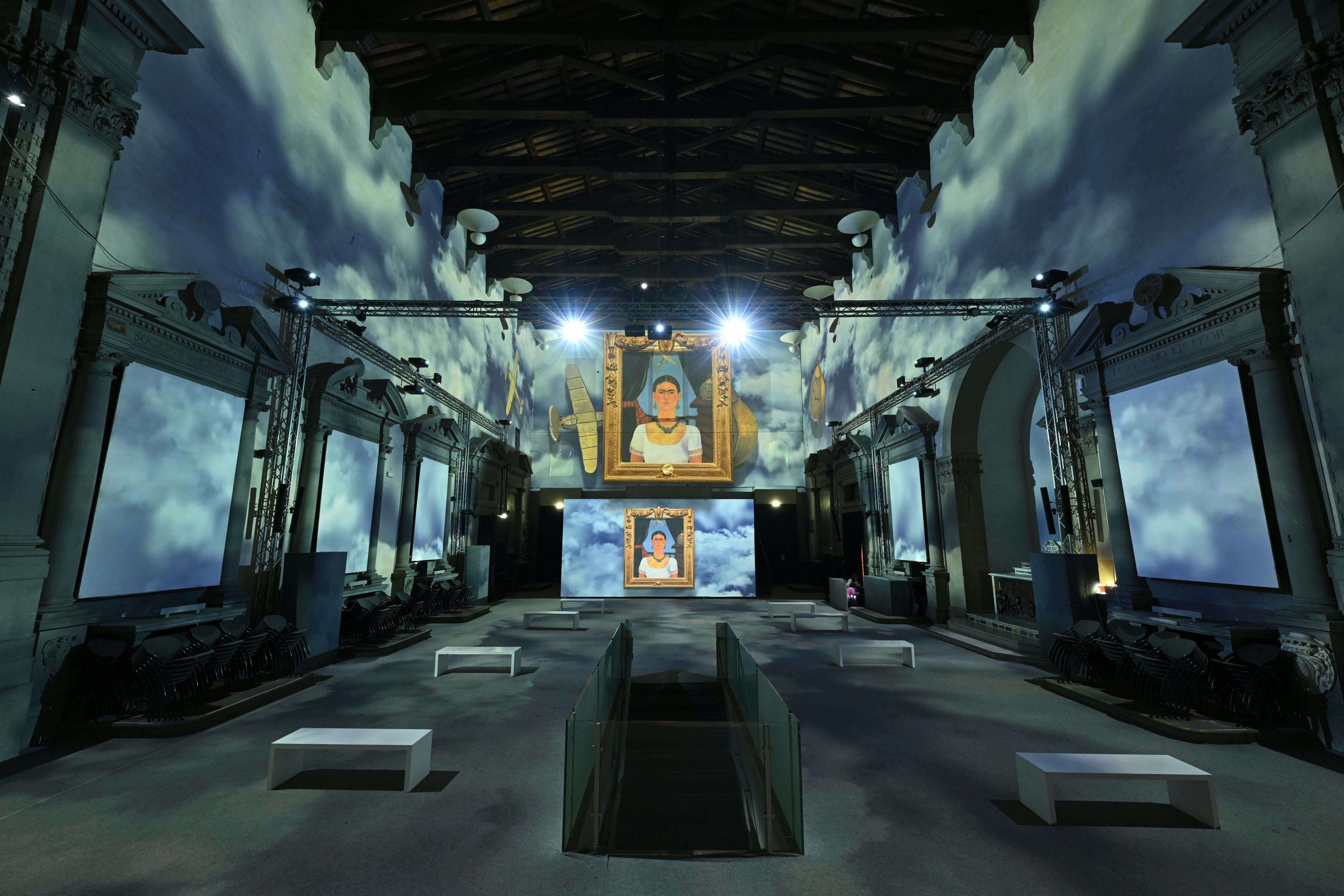 A large art exhibition hall with high ceilings and projected cloud patterns, displaying framed portraits on screens.