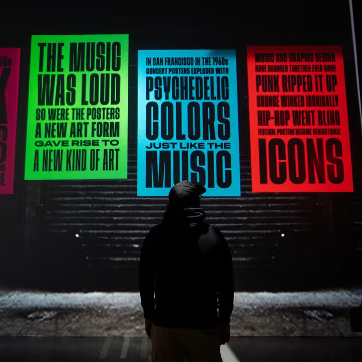 Osoba v tmavé mikině stojí před barevnými plakáty s velkým, odvážným textem o hudbě a umění.