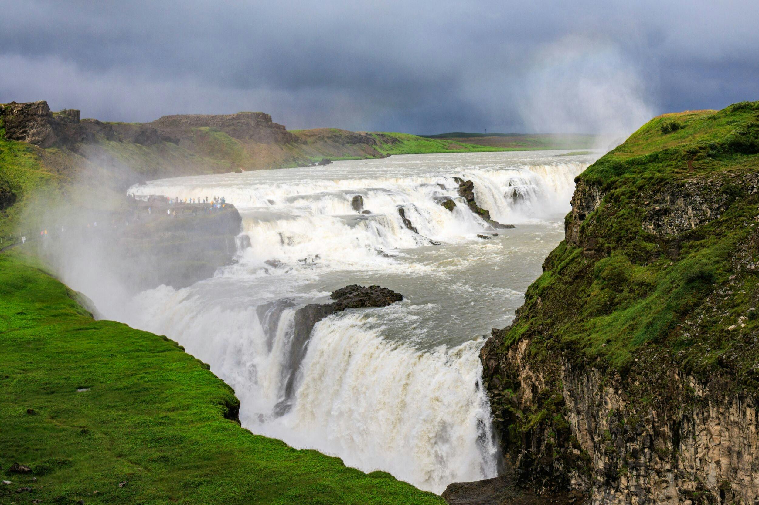 A large waterfall cascading over multiple tiers, surrounded by green cliffs and a cloudy sky.