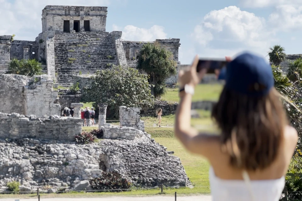 A person photographing an ancient stone ruin with stairs and visitors exploring the site under a blue, partly cloudy sky.