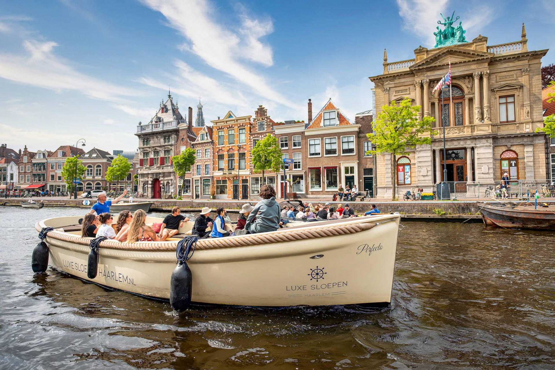 A group of people on a boat in a canal with historic buildings and trees in the background under a partly cloudy sky.
