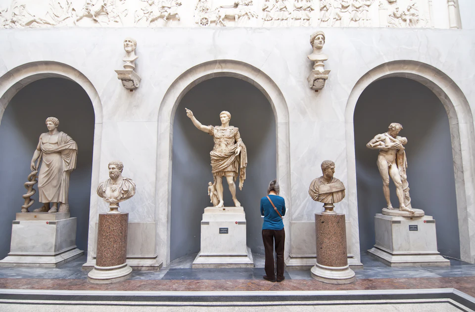 A person standing in front of three marble statues displayed in arched alcoves within a museum setting.