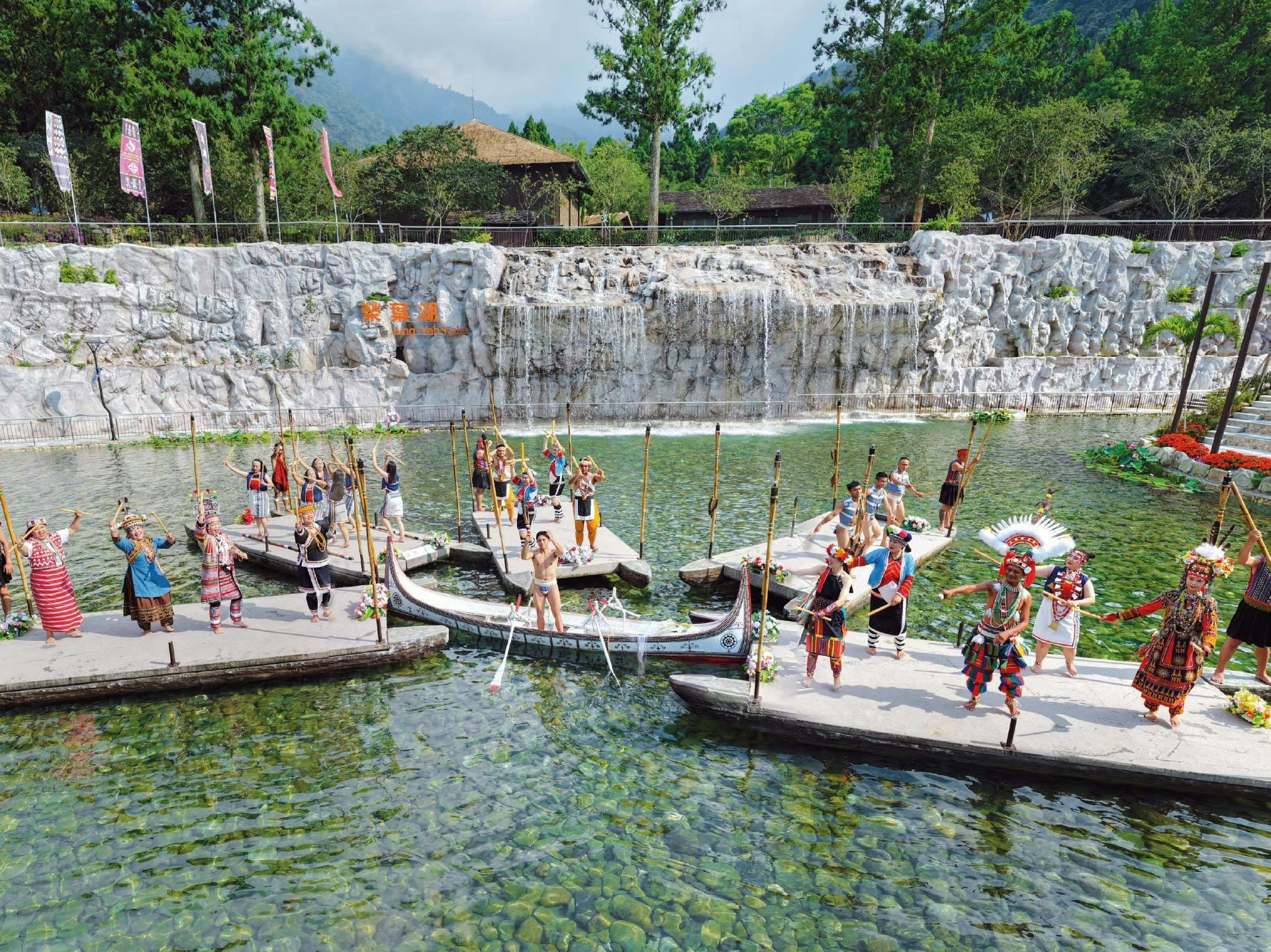 People in traditional attire performing on floating platforms on a clear waterway, with a waterfall and trees in the background.