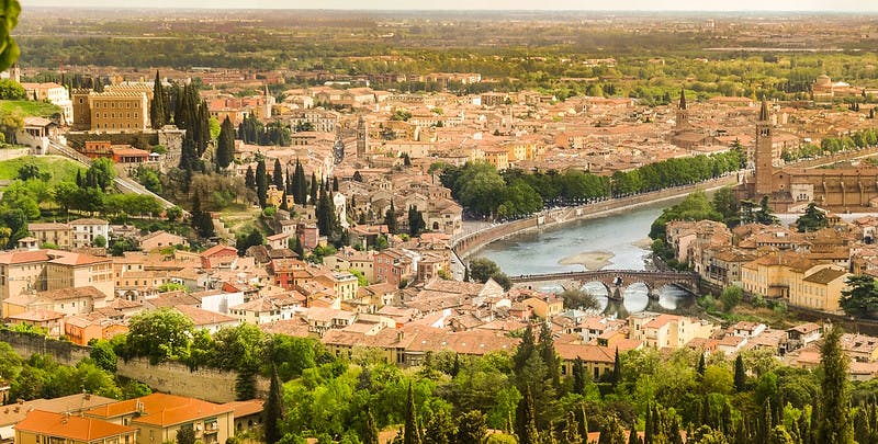 Vista aérea de una ciudad histórica con tejados de tejas rojas, árboles, un río serpenteante y un puente de piedra en medio de un paisaje verde.