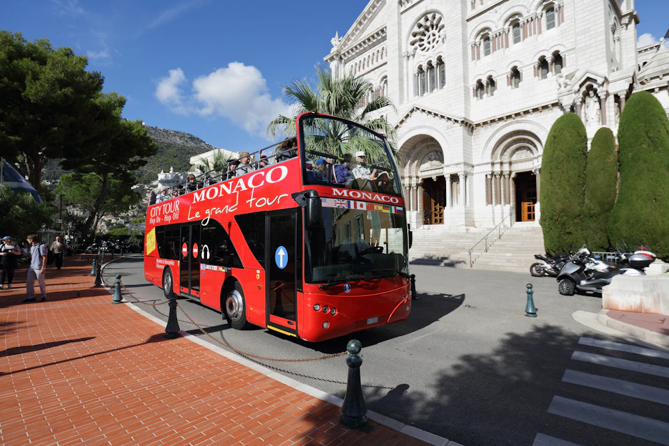 A red double-decker tour bus labeled "Monaco Le grand tour" is parked near a white historic building.
