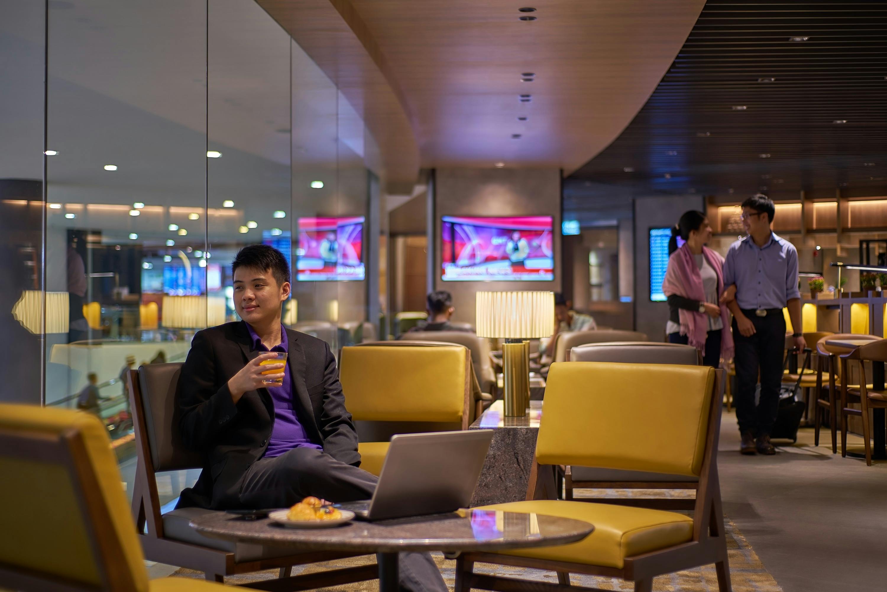 A man in a suit sits at a table in a lounge, holding a drink, with a laptop and a plate of food in front of him.