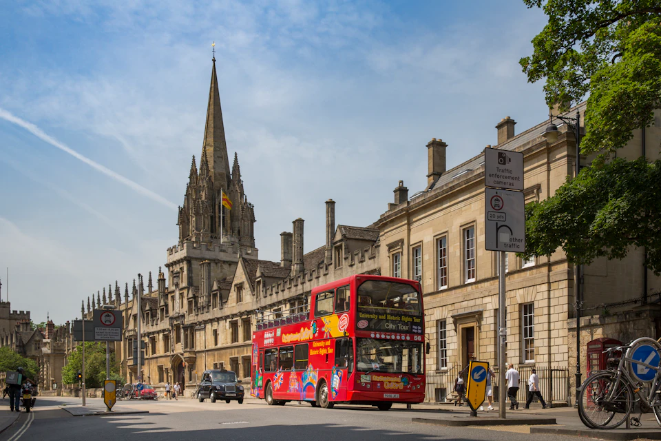 A red double-decker tour bus driving past historic buildings with a church spire on a sunny day.