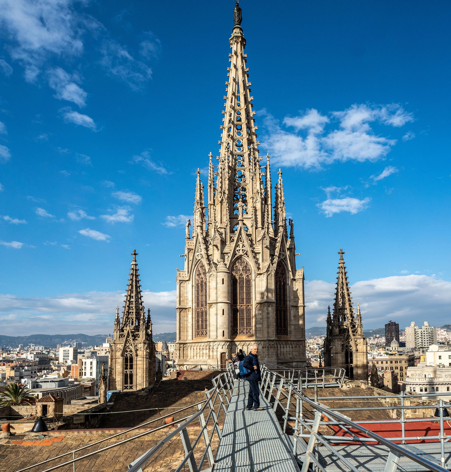 A large Gothic-style cathedral with ornate spires, viewed from a walkway with people on it, under a clear blue sky.