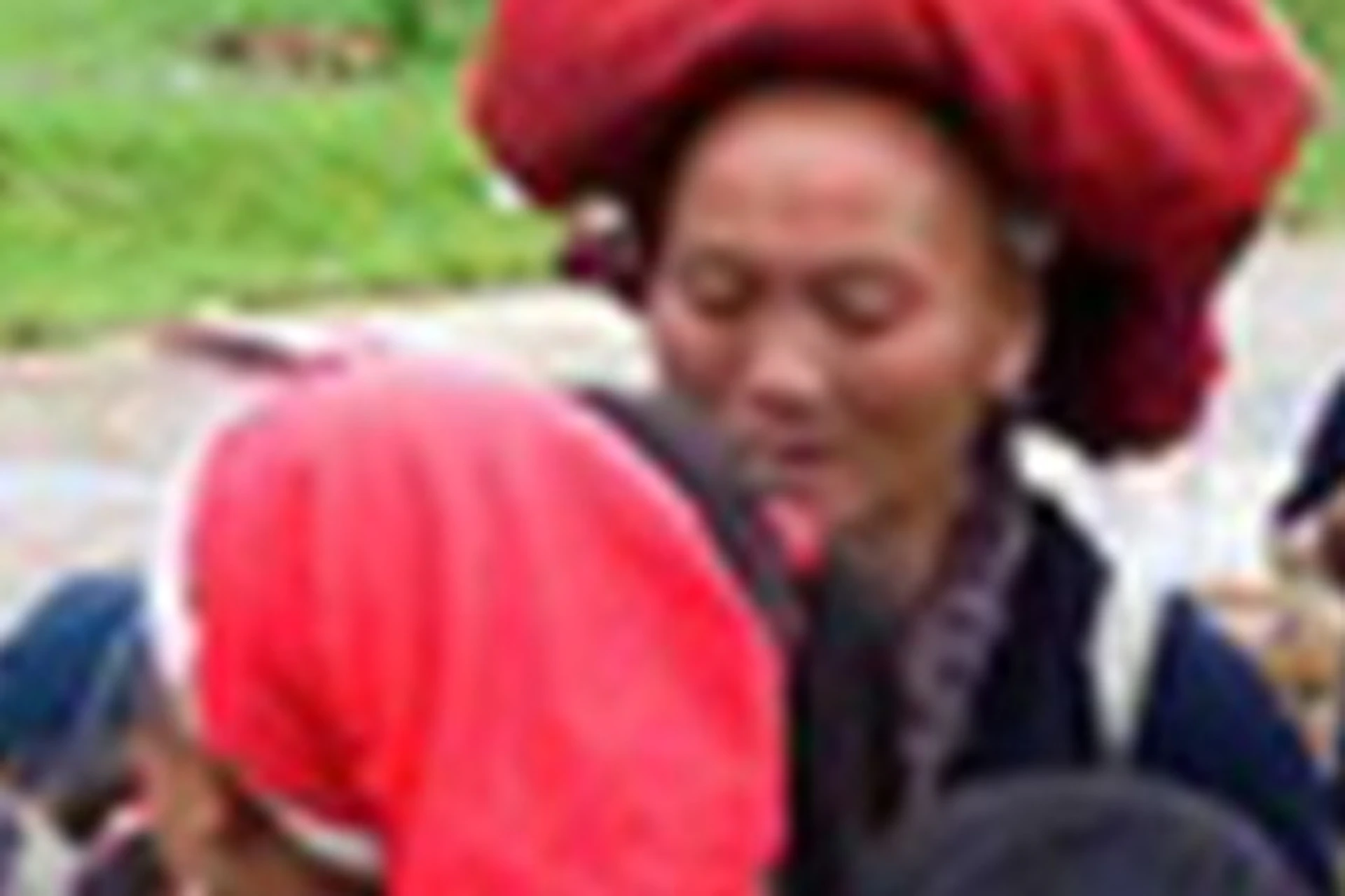 Group of people, mostly women and children, wearing traditional clothing and red headscarves, gathered near wooden buildings and green fields.
