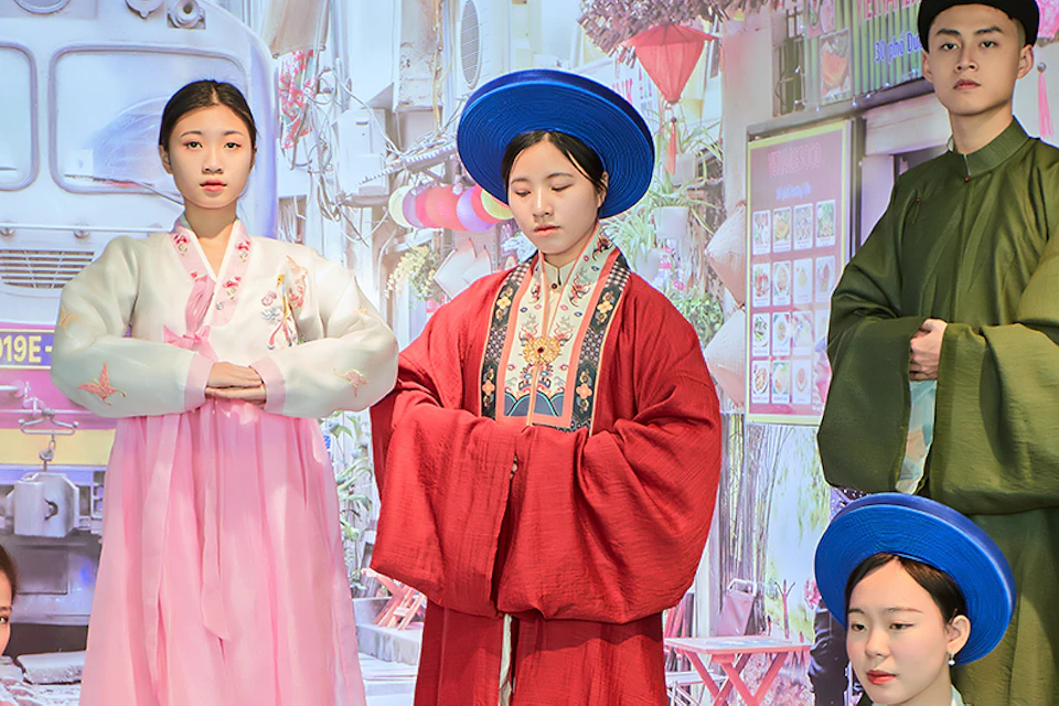 A group of people wearing traditional clothing pose on a stage against a scenic urban background with a train visible.