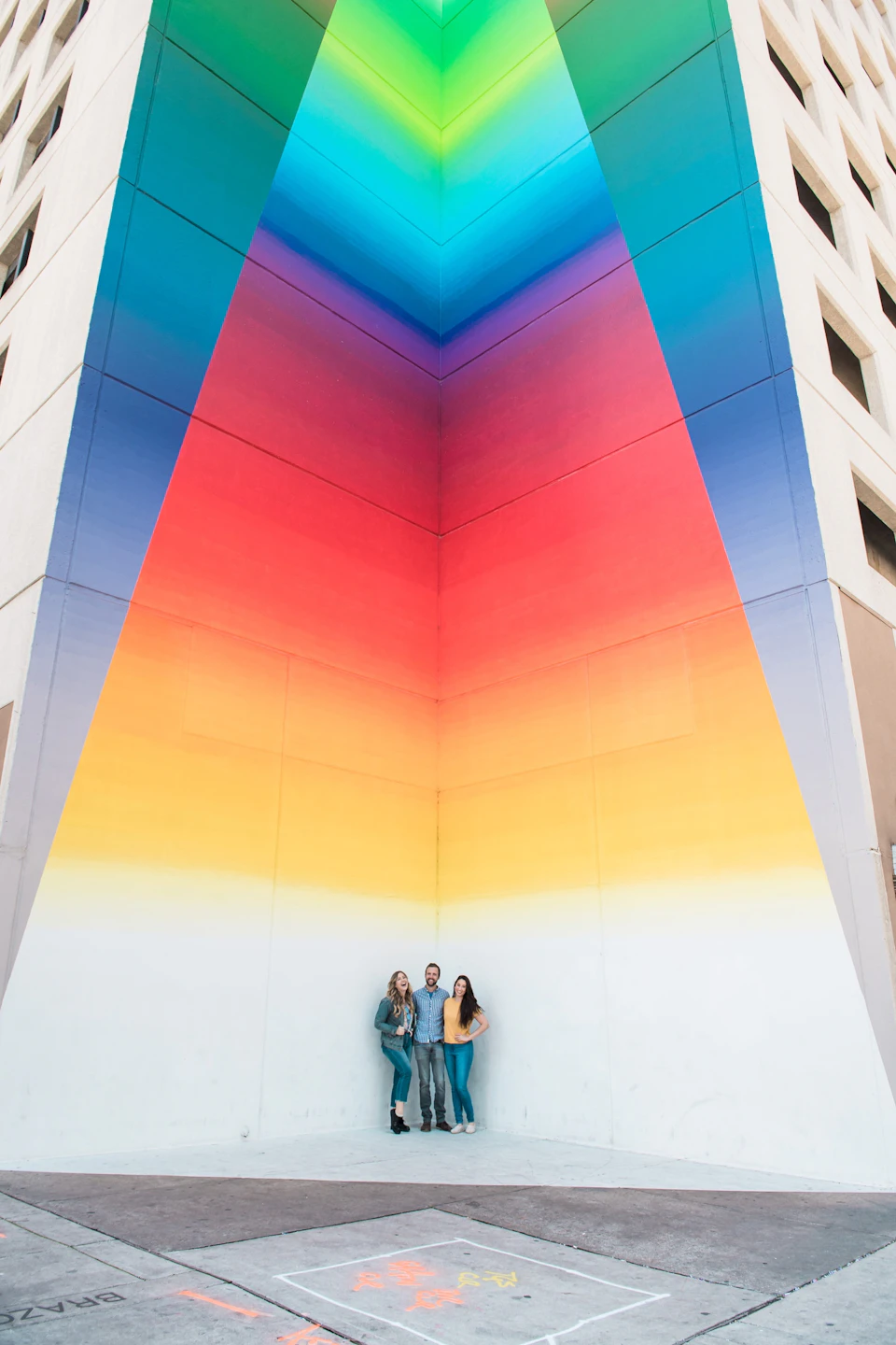 Three people stand against a building wall painted with a colorful gradient design transitioning from blue to red to yellow.