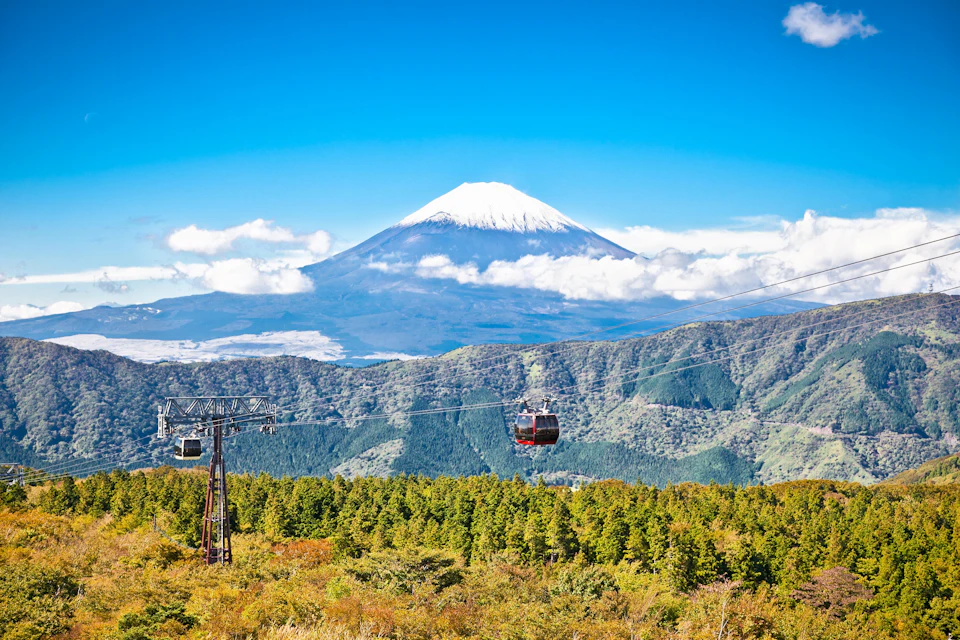 Cable cars suspended over a forest with Mount Fuji in the background under a clear blue sky.