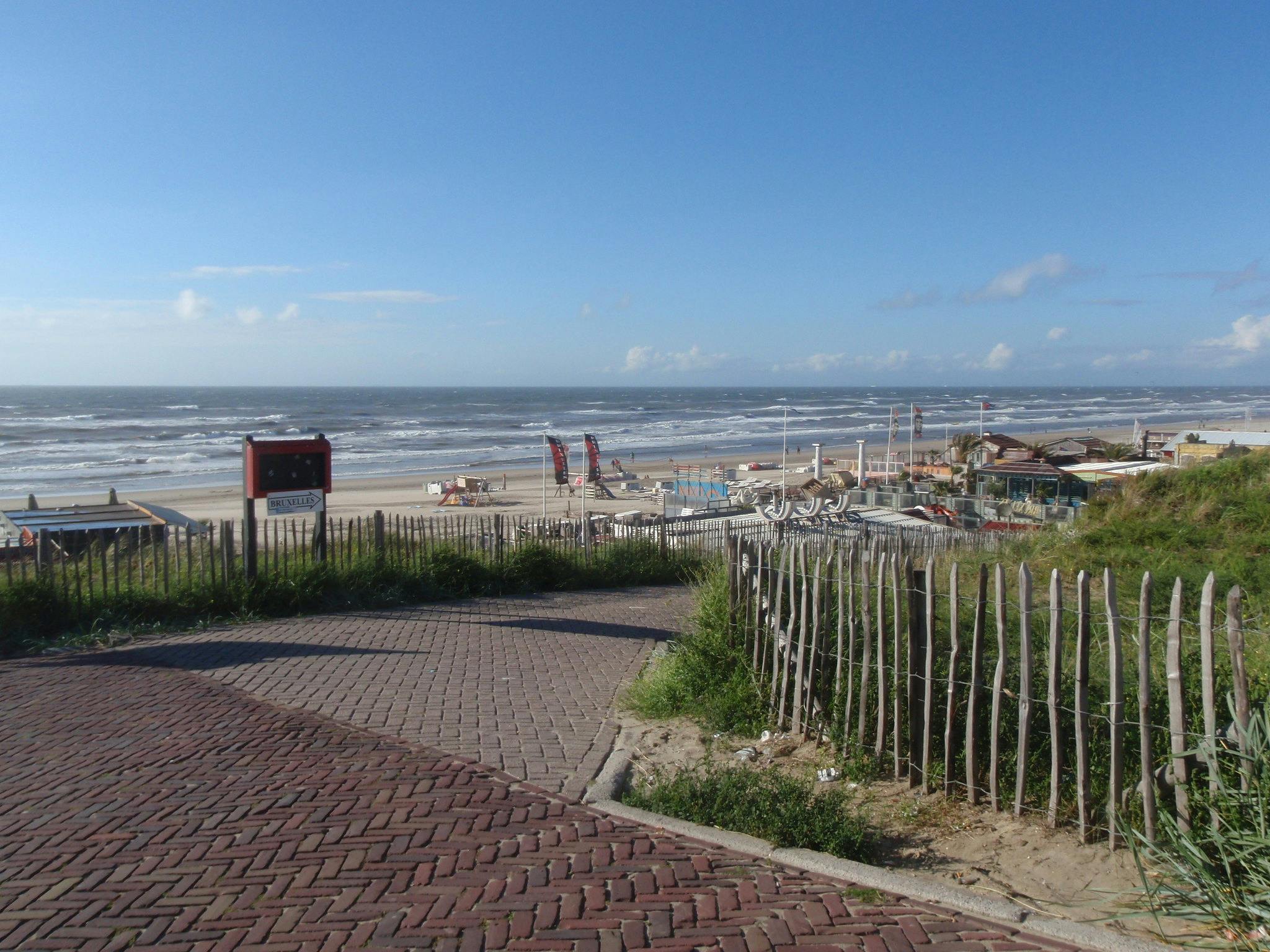 포장된 길은 파도, 해변 의자, 몇 개의 건물이 있는 해변으로 이어집니다. 나무 펜싱과 잔디가 길과 경계를 이룹니다. 위쪽의 푸른 하늘.