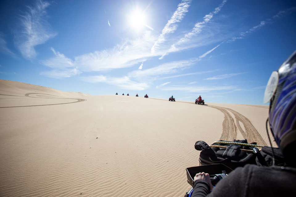 People riding ATVs on sandy dunes under a bright sun with clear skies; tire tracks visible in the sand.