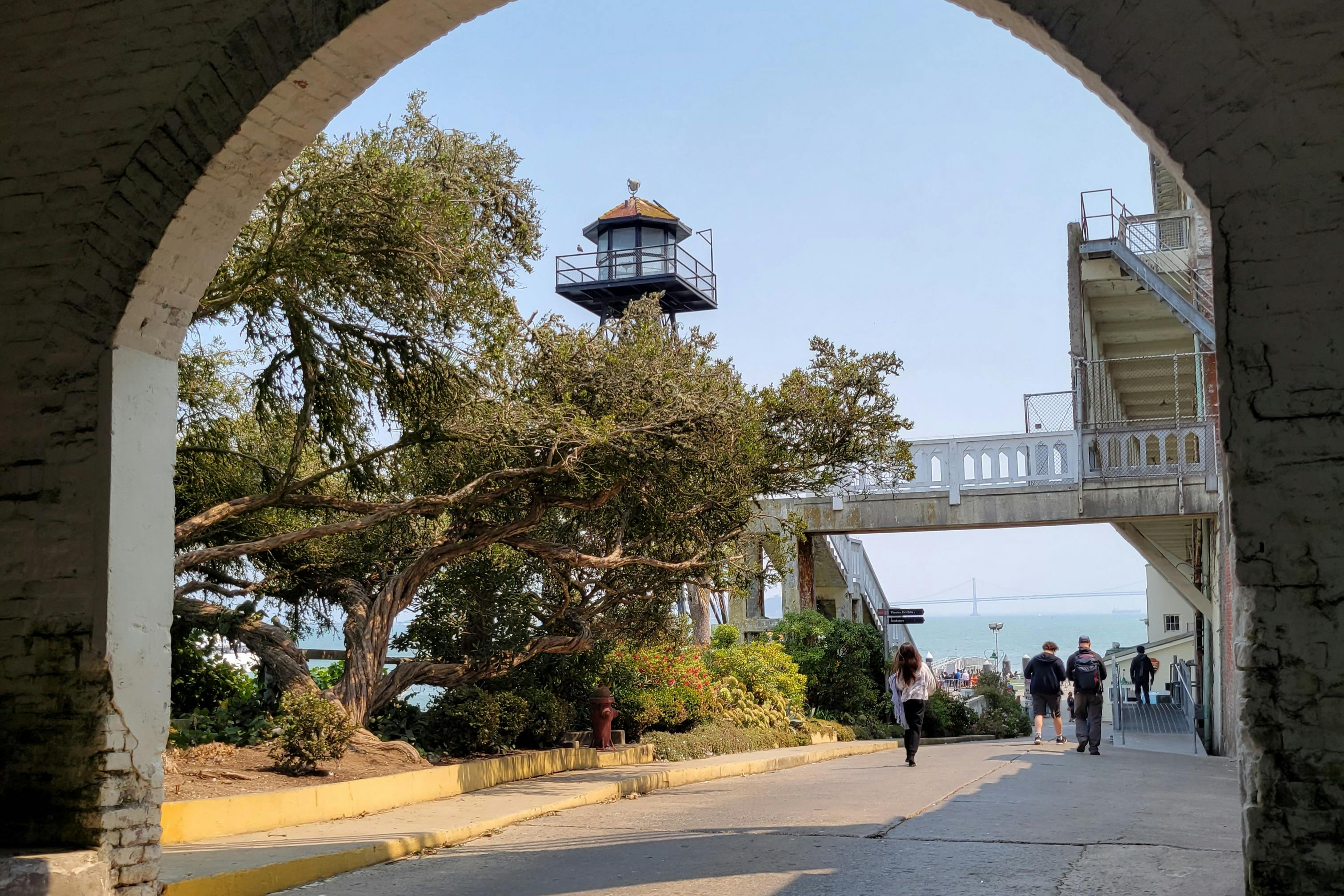 A lighthouse and trees viewed through an archway, with a walkway and people in the foreground.