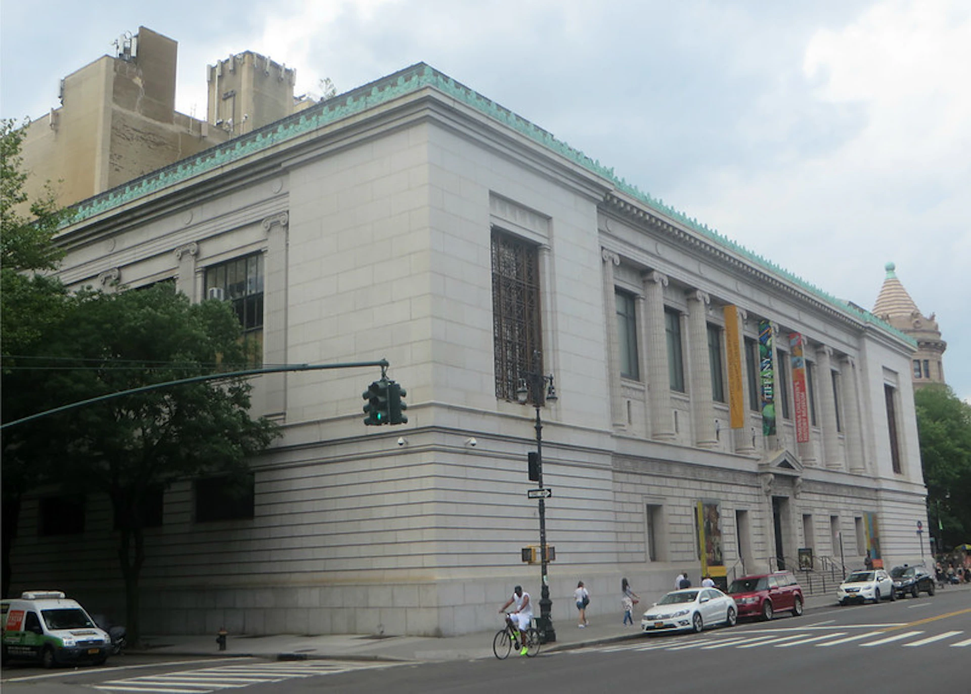 Historic building on a city corner with banners hanging, pedestrians, a bicyclist, and parked cars in the foreground.