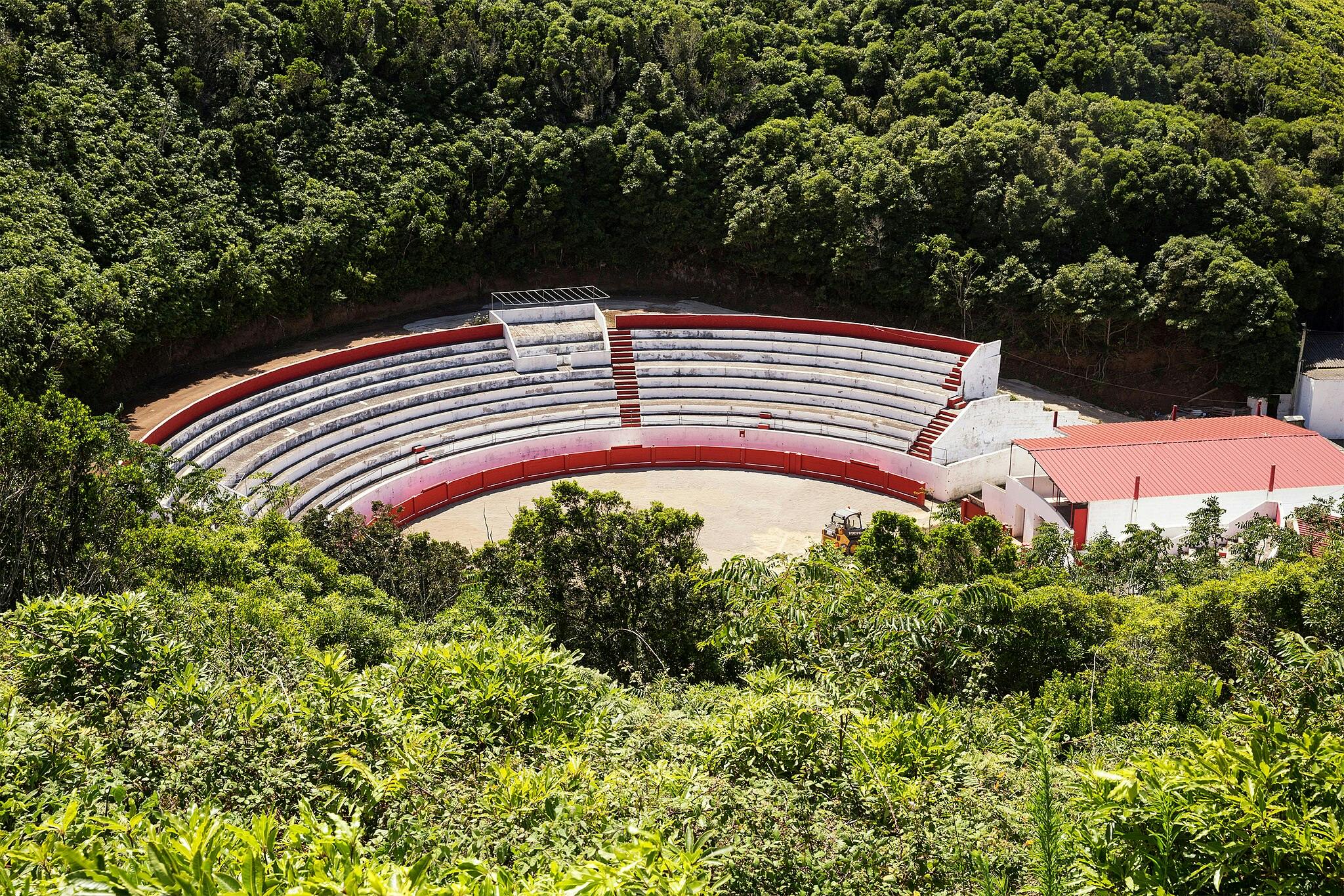 Von oben sieht man ein leeres Amphitheater mit rot-weißer Bestuhlung, umgeben von einem dichten grünen Wald.
