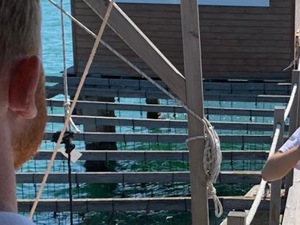 A guide speaks to two people on a wooden platform over water with a sign reading "Musclarium" in the background.