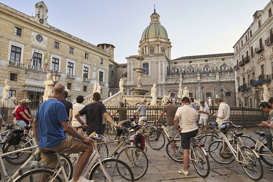 A group of cyclists gathered in front of a historic fountain and ornate building with statues and a domed structure in the background.