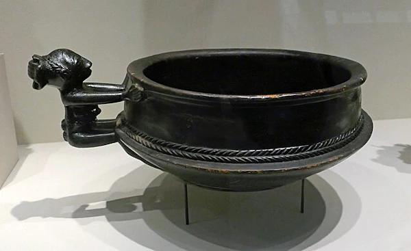 A black, antique bowl with intricate braided designs and a handle on a display stand.