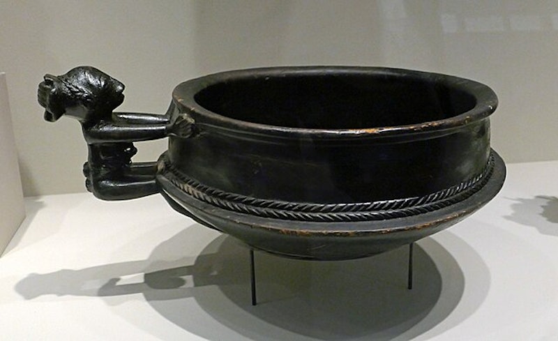 A black, antique bowl with intricate braided designs and a handle on a display stand.