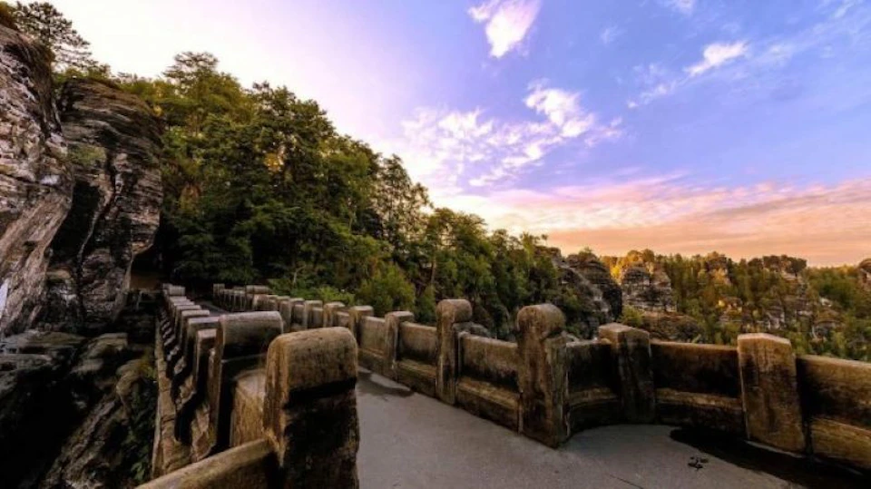 Stone walkway with guardrails curving alongside dense trees at sunrise or sunset, with a colorful sky and rugged landscape in the distance.