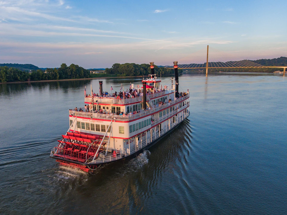 A paddle steamboat filled with passengers sails on a calm river, with trees and a distant bridge in the background.