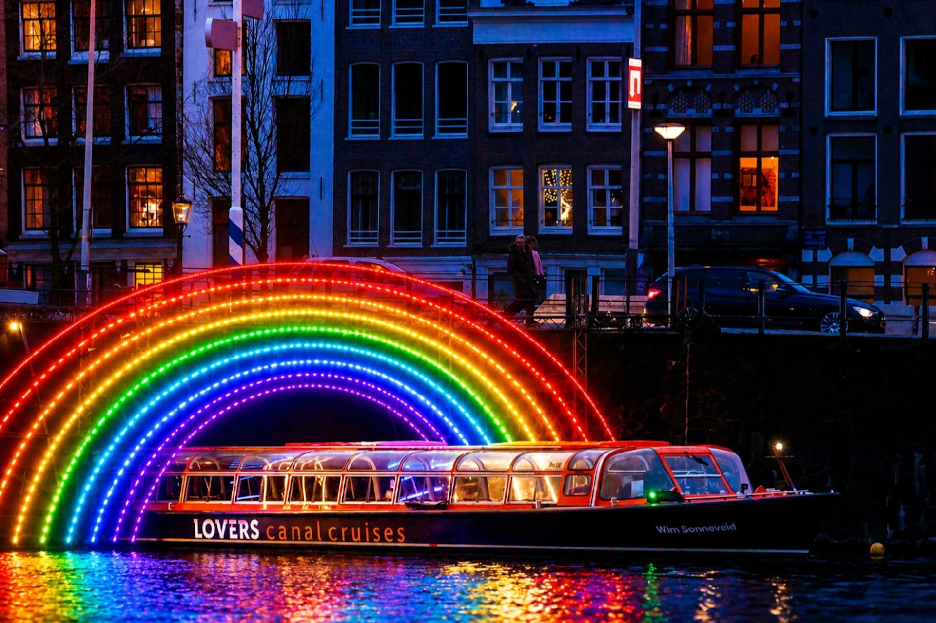 A canal cruise boat passing under a brightly lit rainbow arch at night, with buildings in the background.