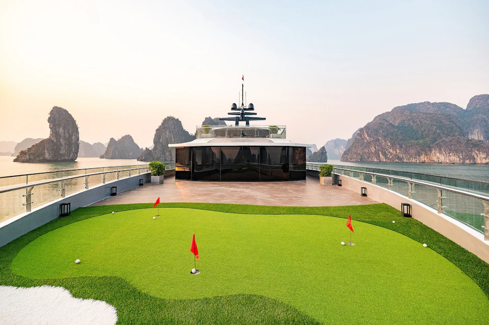 A yacht's deck features a mini golf course with red flags, lounge chairs, potted plants, and scenic limestone cliffs in the background.