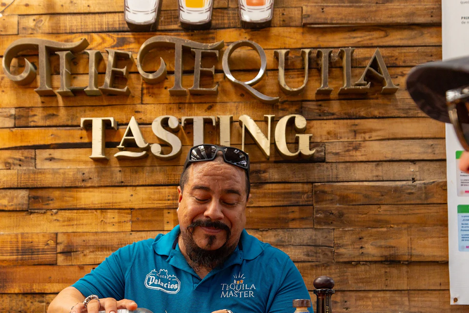 A man pours tequila into a small cup at a bar with a wooden backdrop labeled "The Tequila Tasting." Two people watch.