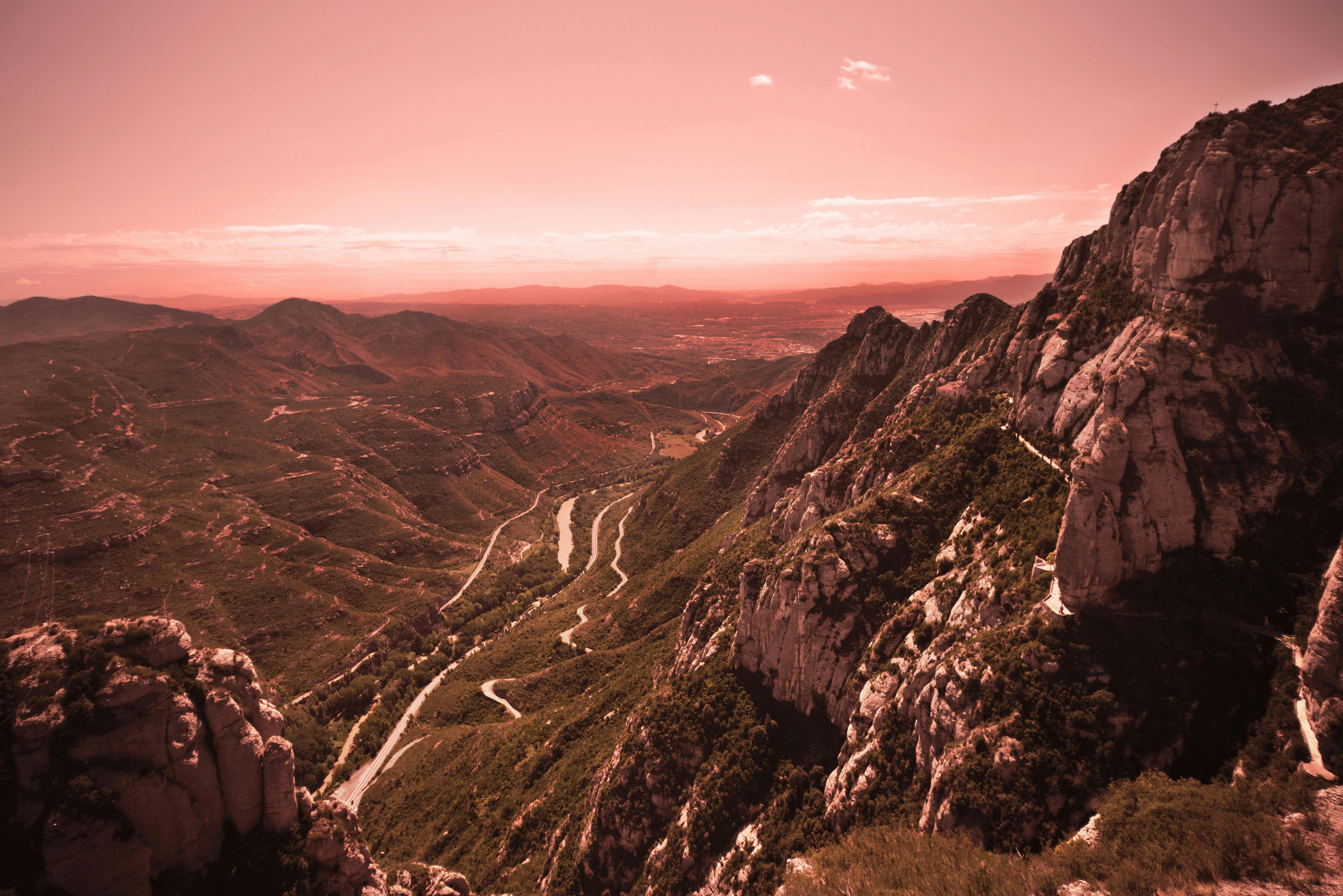 A winding road cuts through a green mountain valley under a pink-tinted sky. Rocky cliffs are visible in the foreground.