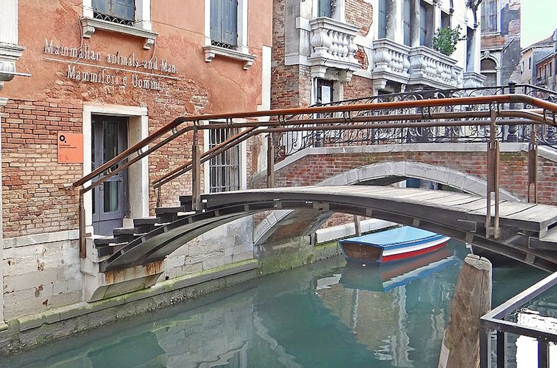 An arched wooden bridge spans over a narrow canal in Venice, with a blue and red boat moored in the water below.