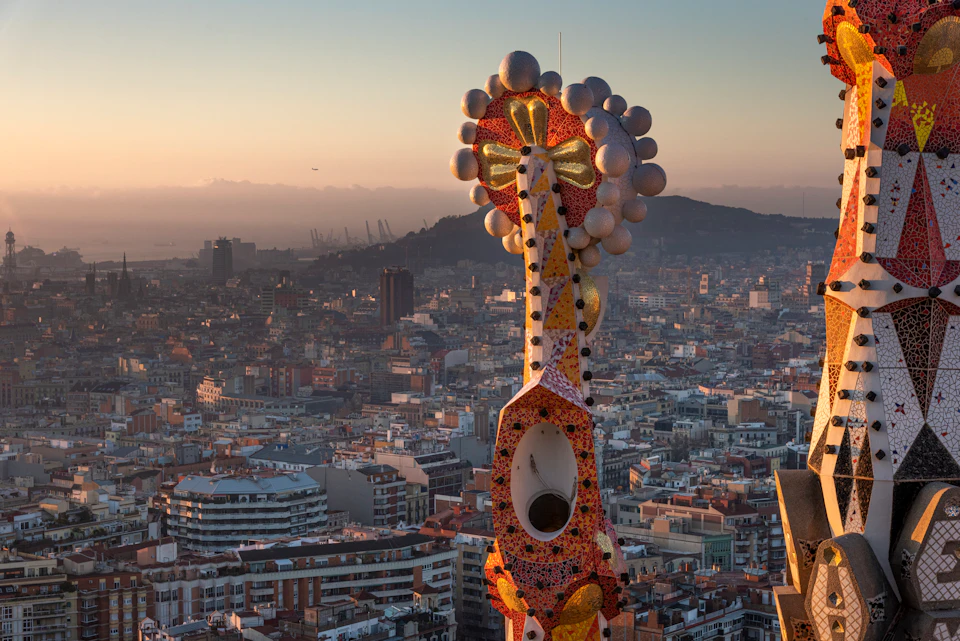 A colorful, mosaic tower with circular ornaments overlooks a city skyline at sunset.