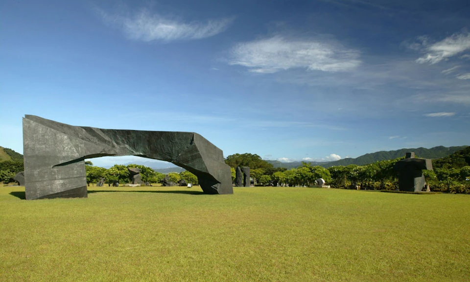 A large black sculpture on a grassy field with trees and mountains in the background under a clear blue sky.