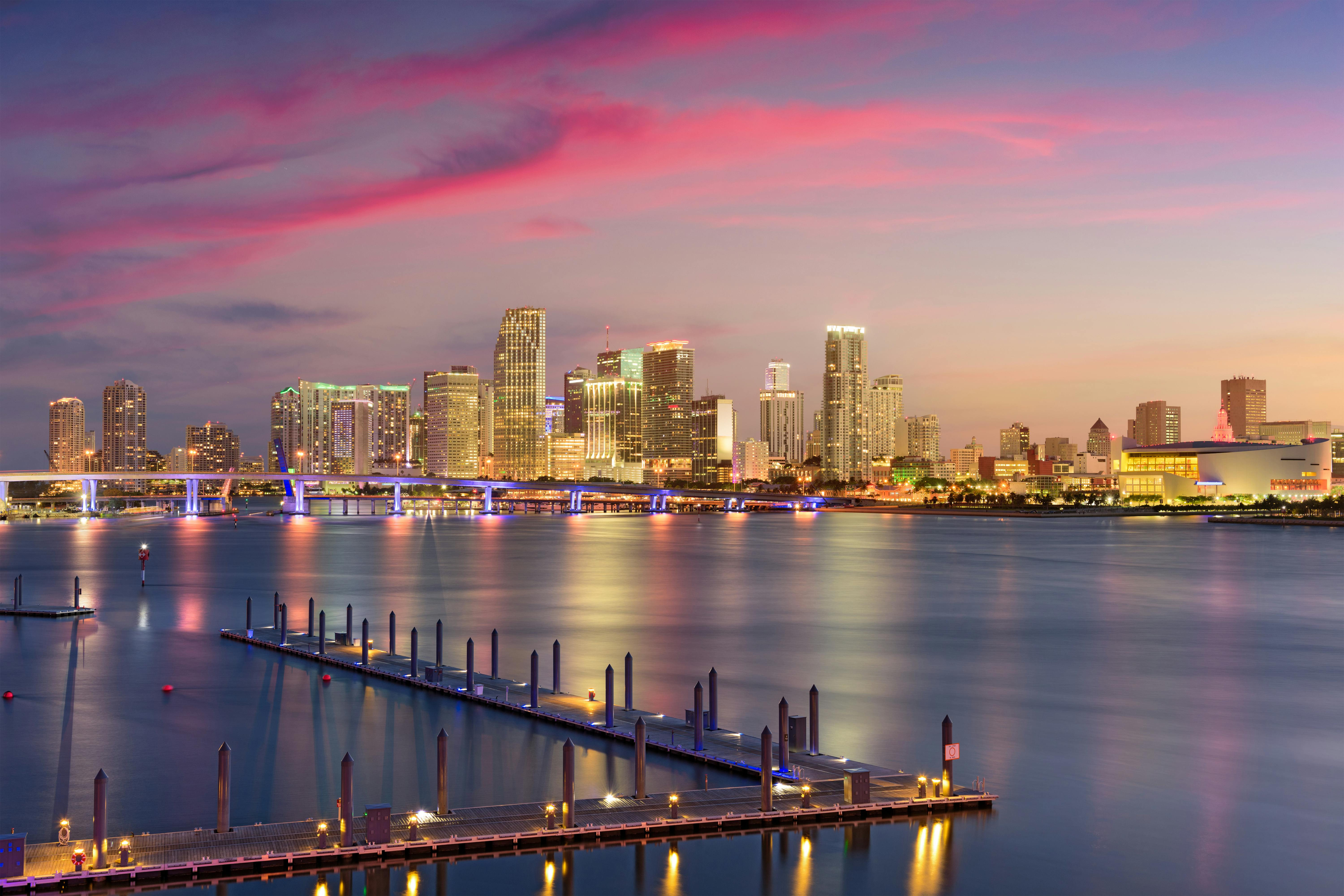 A city skyline illuminated at dusk with vibrant pink clouds overhead, a lit-up bridge, and a calm harbor with empty docks.