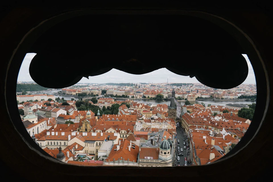 View of a cityscape with red-roofed buildings, a river in the background, and streets lined with cars, seen through an archway.
