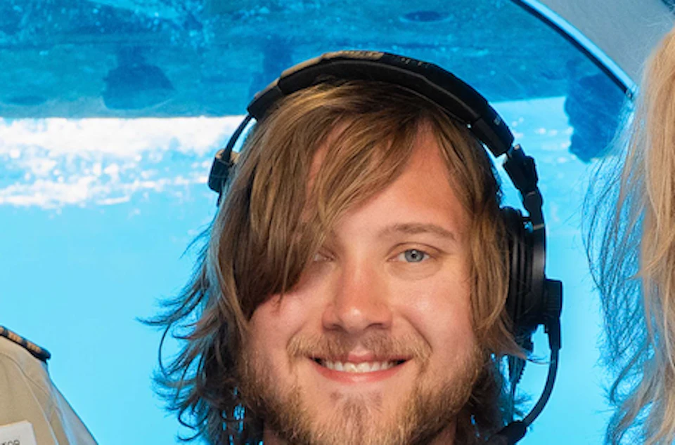 Three people sitting inside a blue submarine; one man wearing headphones, another smiling, and a woman in a sleeveless top.