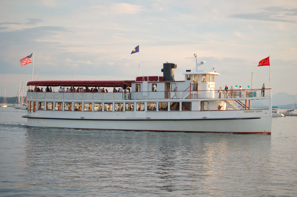 A white ferry with a red roof deck and a flag, carrying passengers, sailing on calm water under a partly cloudy sky.