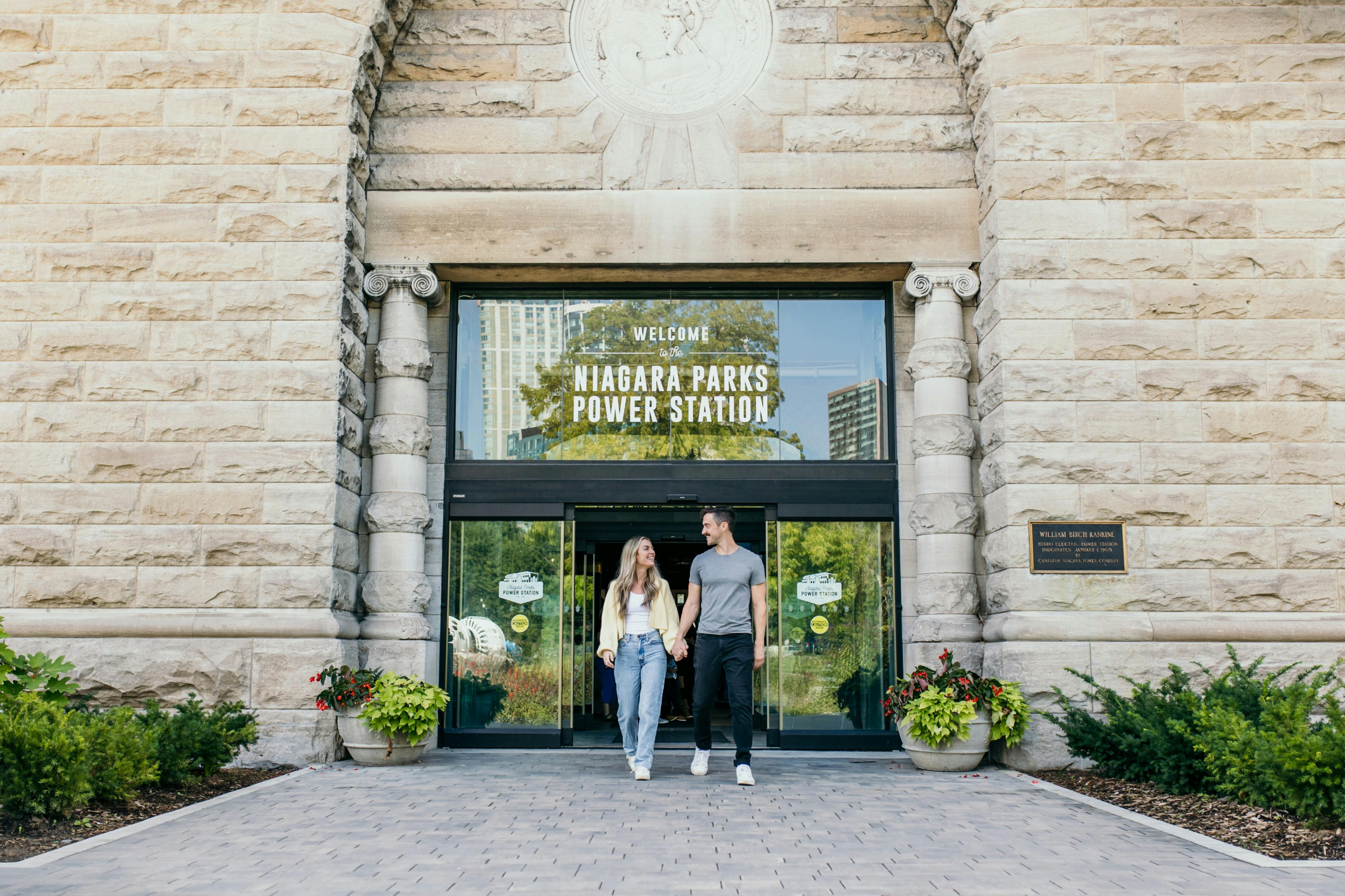 나이아가라 공원 발전소 입구 앞에서 손을 잡고 있는 두 사람은 석조 건축물과 식물로 둘러싸여 있습니다.