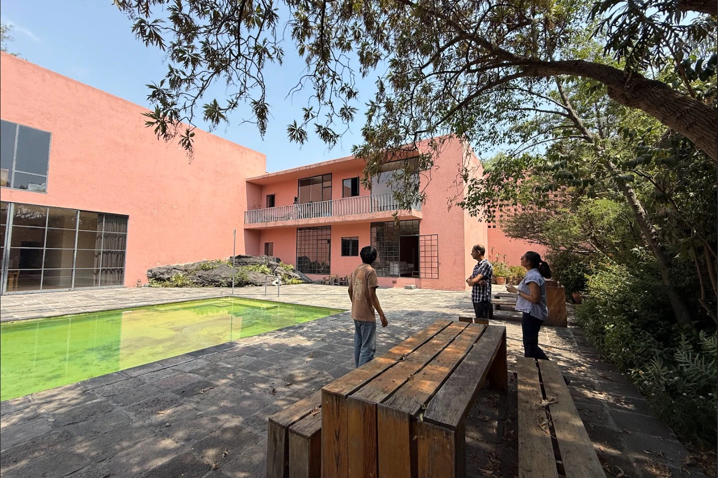 Three people stand in a courtyard with a pink building, a pool, wooden benches, and trees under a clear sky.