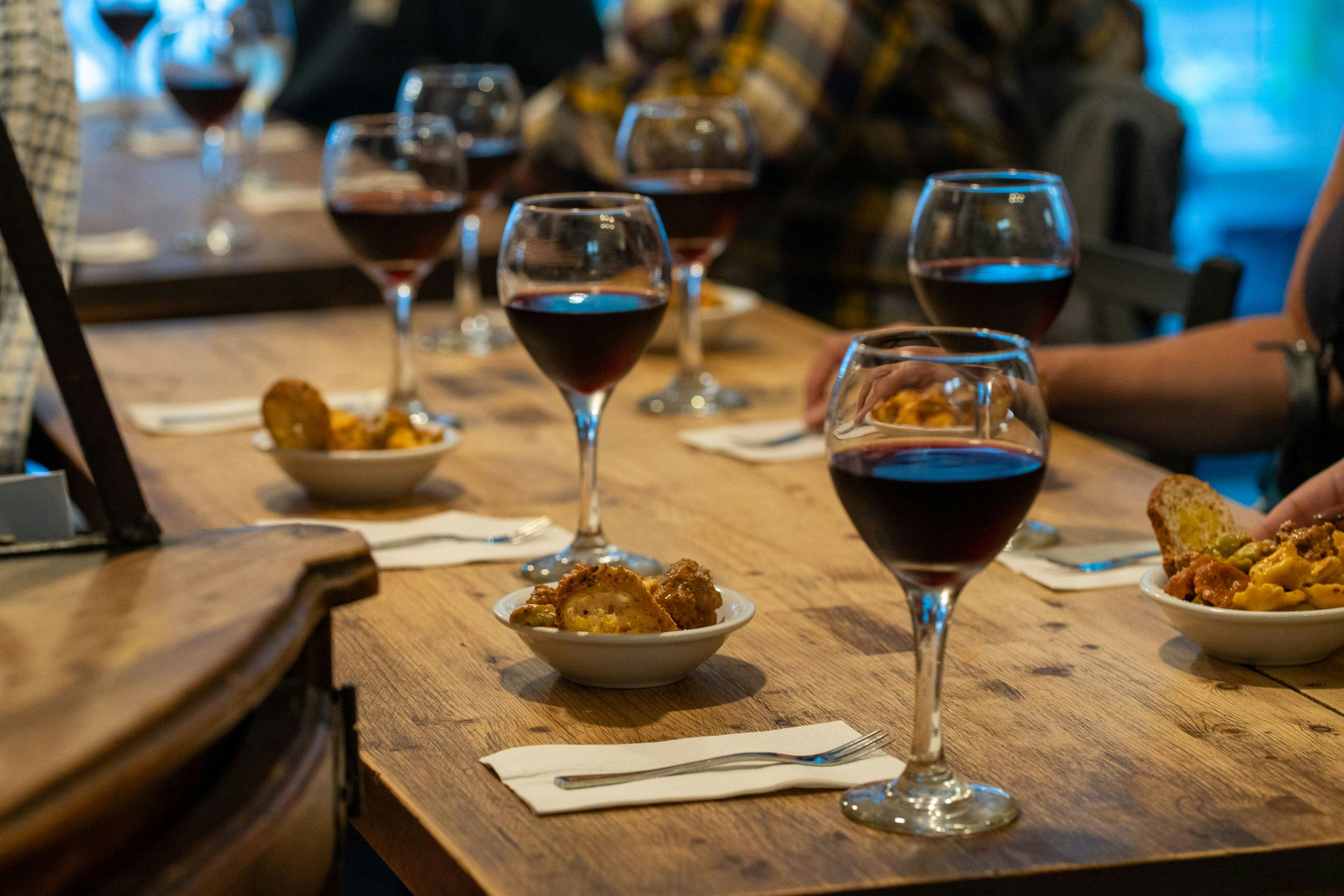 Kilka kieliszków czerwonego wina i małe miseczki przystawek na drewnianym stole z serwetkami i widelcami; rozmazani ludzie w tle.