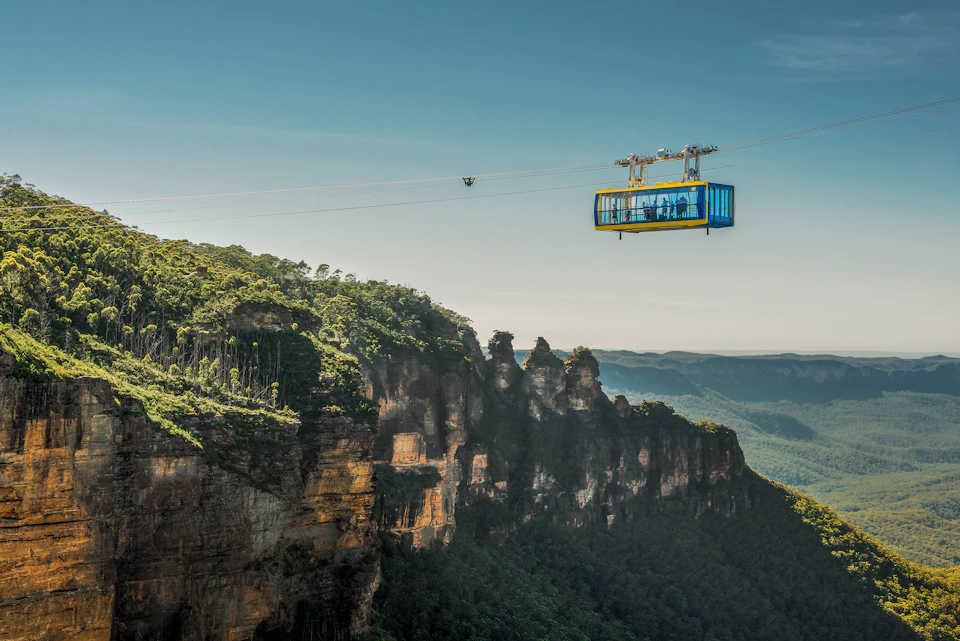 A yellow and blue cable car suspended above a forested cliff with distant mountains and a clear sky in the background.