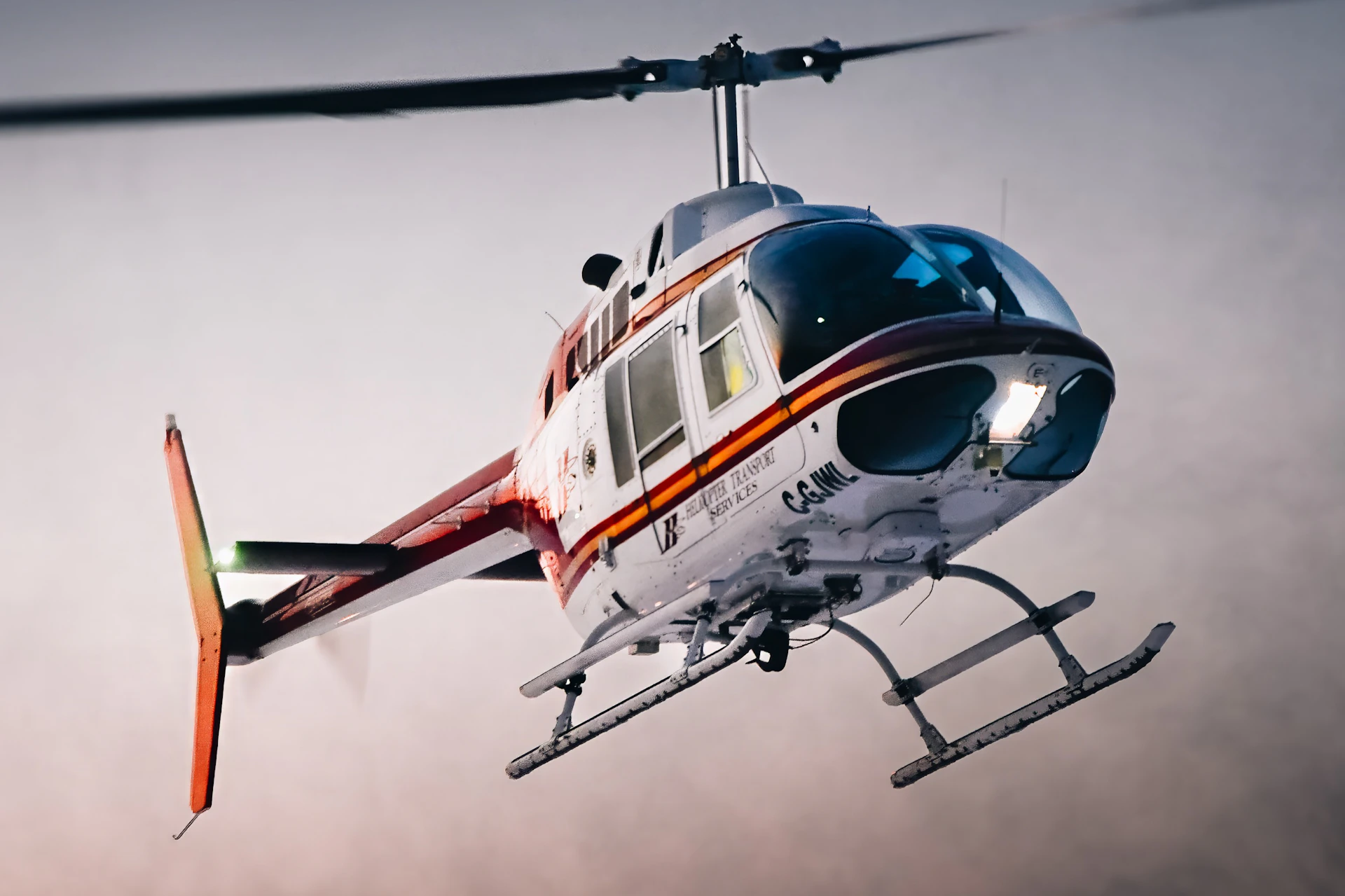 White and red helicopter in mid-flight with visible landing skids and rotating blades against a cloudy sky.