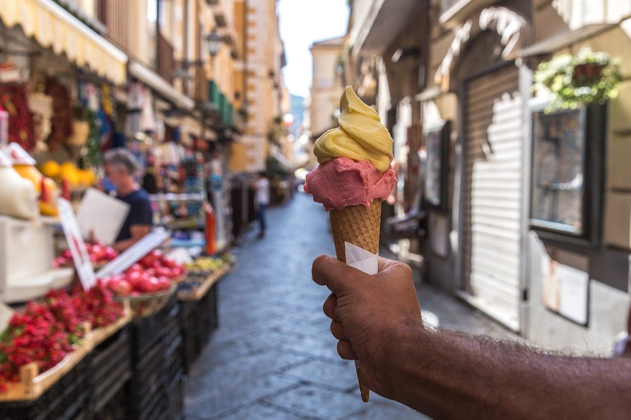 Een hand die een hoorntje met geel en roze ijs vasthoudt in een smalle, drukke straat met winkels en mensen op de achtergrond.