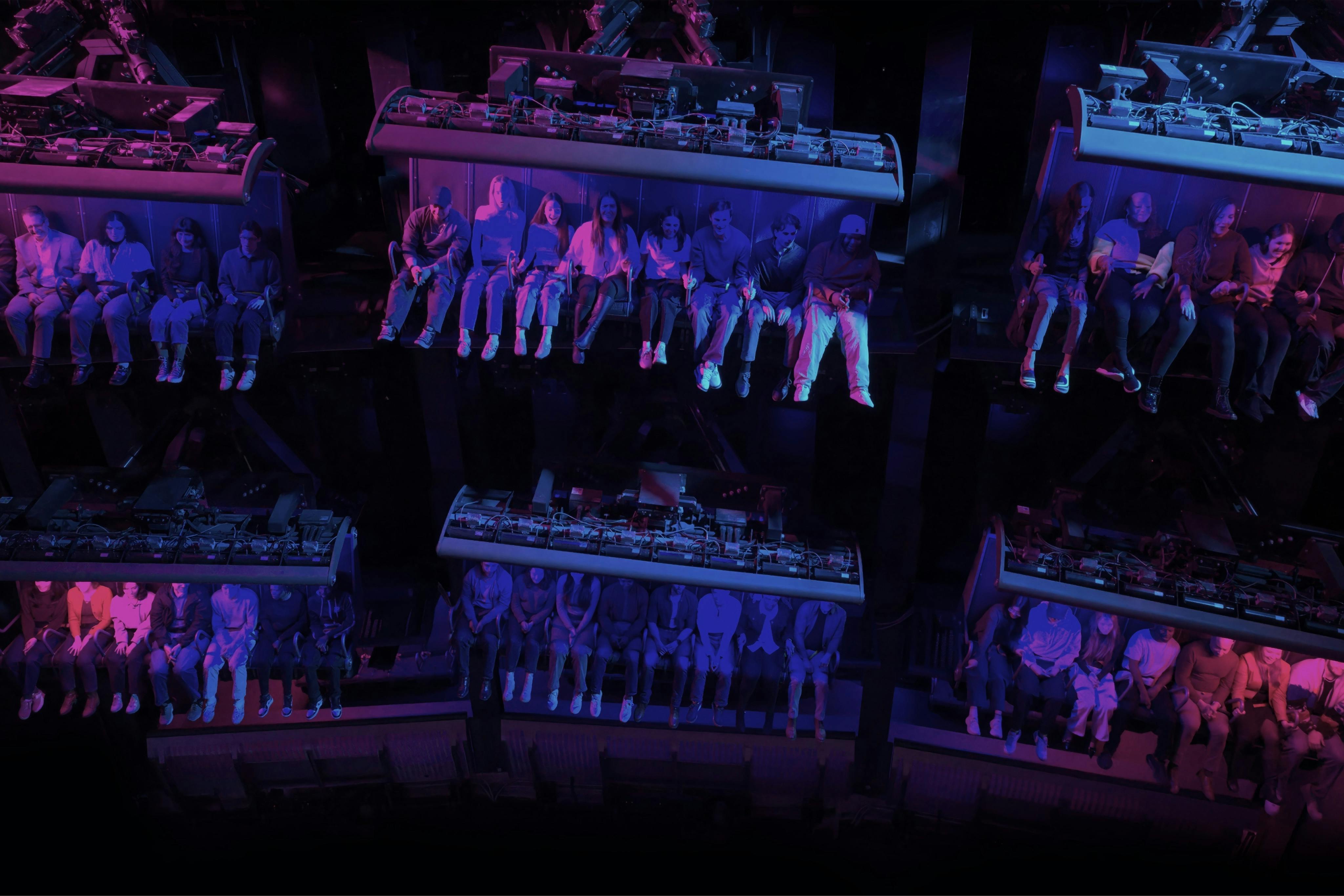 Reihen von Menschen, die in einem dunklen Fahrgeschäft sitzen, beleuchtet von violetten und blauen Lichtern.