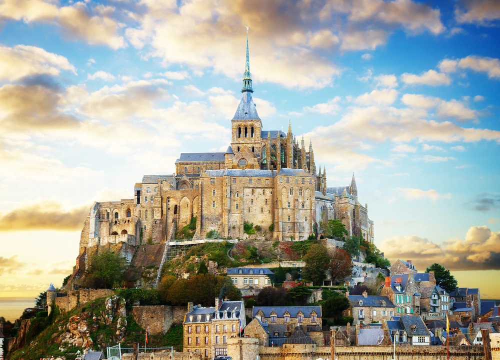 A historic stone abbey with a tall spire situated on a hill, surrounded by smaller buildings under a partly cloudy sky.