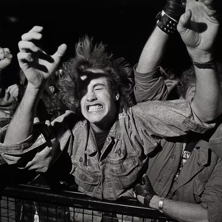 Člověk v džínové bundě, se zavřenýma očima, svírající bariéru na přeplněné akci s rukama zvednutýma kolem sebe.