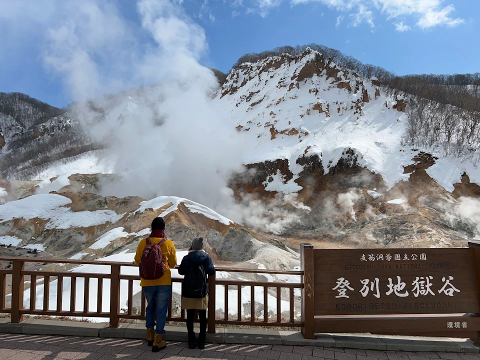 Two people stand at a wooden railing overlooking a steaming, snow-covered volcanic landscape under a partly cloudy sky.